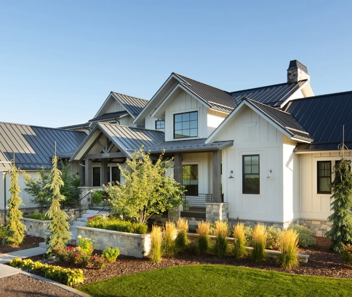 White farmhouse with a green lawn, ornamental grasses, young trees, and layered planting beds